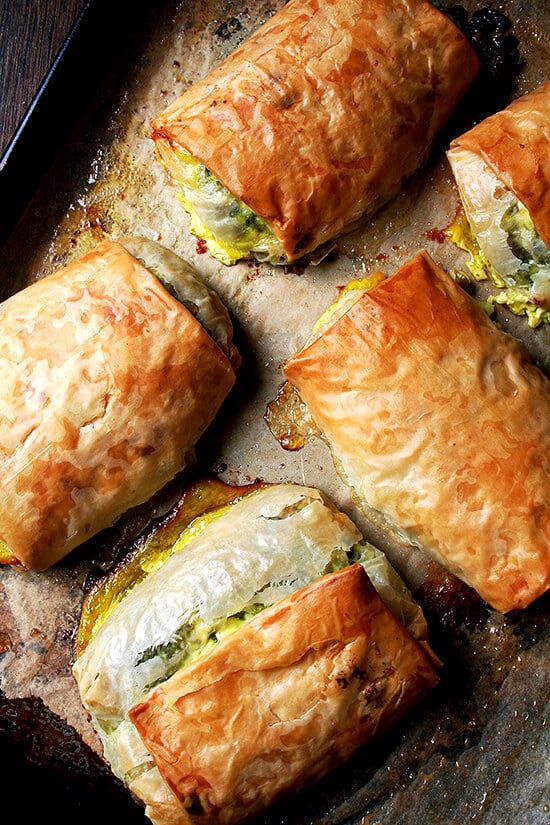 When baked in strudel form, spanakopita assumes an almost breakfast croissant-like character, a perfect bundle of flaky pastry, egg, cheese, and greens. Yum. // alexandracooks.com Five baked spanakopita strudels on a sheet pan.