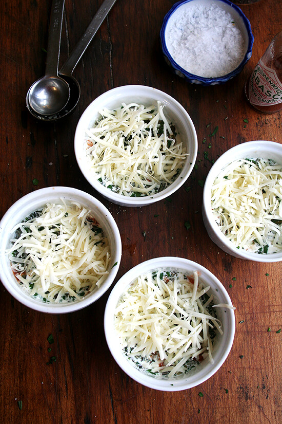 4 ramekins each holding an egg, cheese and herbs, ready for the oven.
