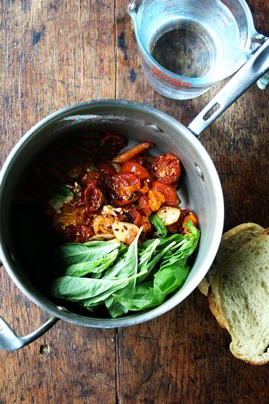 A saucepan filled with roasted vegetables and basil aside a liquid measure with water and a few slices of stale bread.