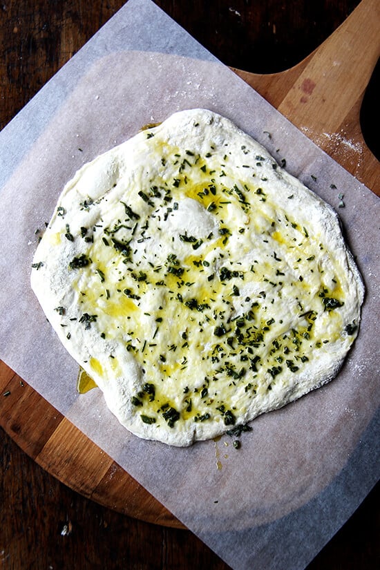 An herbed unbaked flatbread on a sheet of parchment on a baking peel.