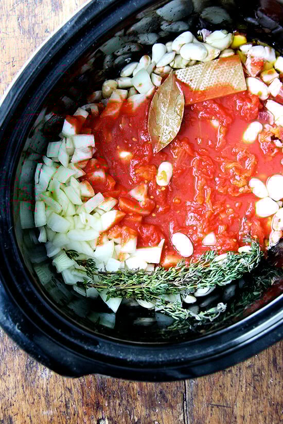 A crockpot filled with onions, beans, pancetta, bay leave, thyme, a parmesan rind, and crushed tomatoes.