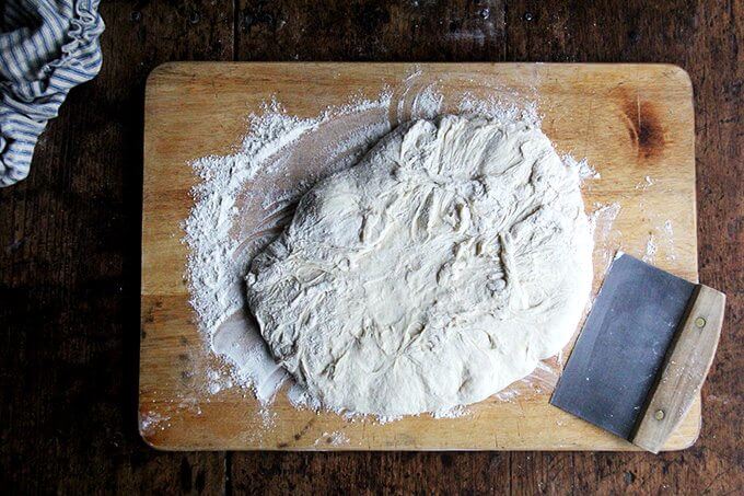 Pizza dough on a board with flour and a bench scraper.