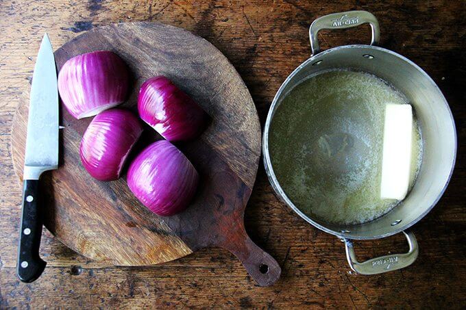 A board of halved onions aside a pot with a stick of butter in it.