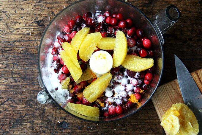 A food processor filled with cranberries, sugar, orange zest, and orange slices.