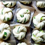 A board with 12 shaped Chinese scallion buns, each on a square of parchment paper.