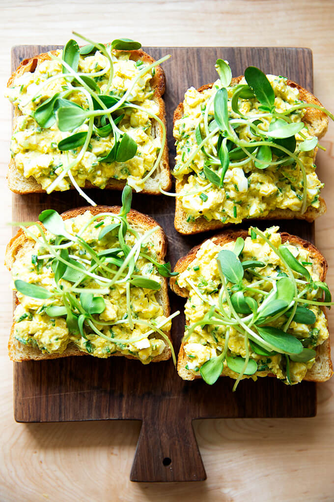 Four slices of toasted sourdough bread topped with avocado-egg salad and sprouts.