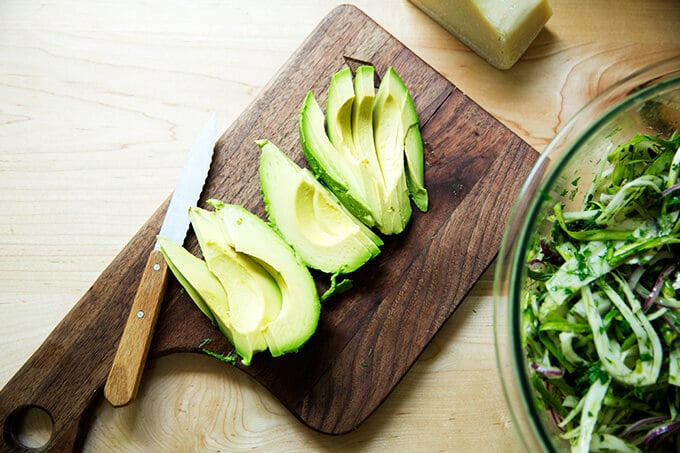 A cutting board with avocado, sliced for fennel salad.