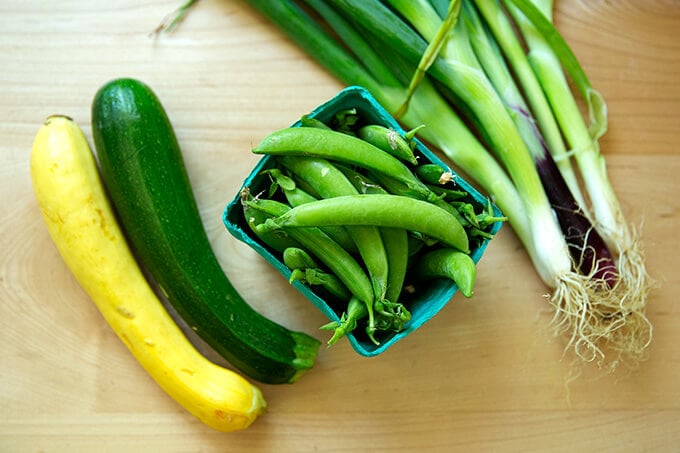 A cutting board with summer squash, snap peas, and spring onions.