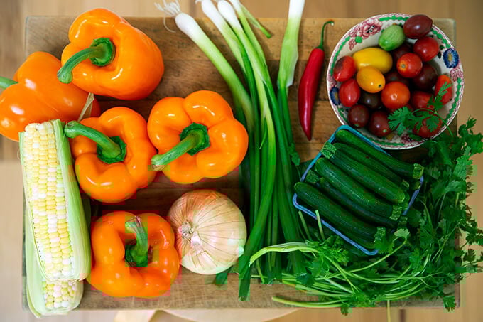 A board with vegetables: orange bell peppers, onion, scallions, zucchini, tomatoes, scallions, and cilantro. 
