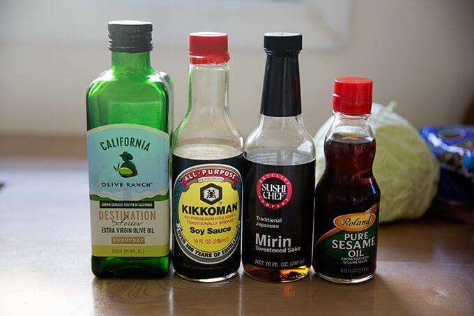 Four bottles standing in a row on a countertop: olive oil, soy sauce, mirin, and sesame oil.