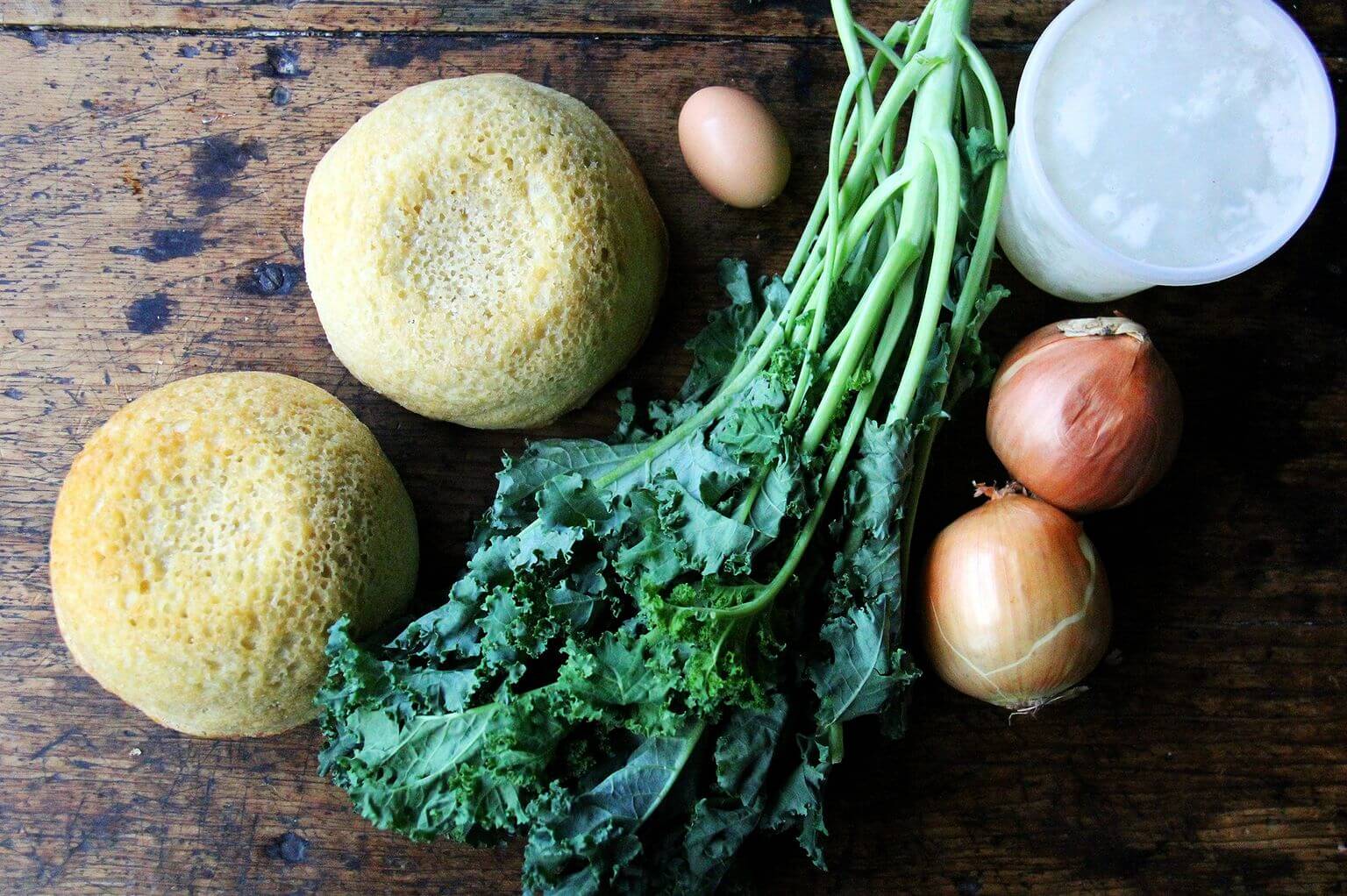 An overhead shot of two loaves of peasant bread, kale, onions, an egg, and chicken stock: ingredients for stuffing.