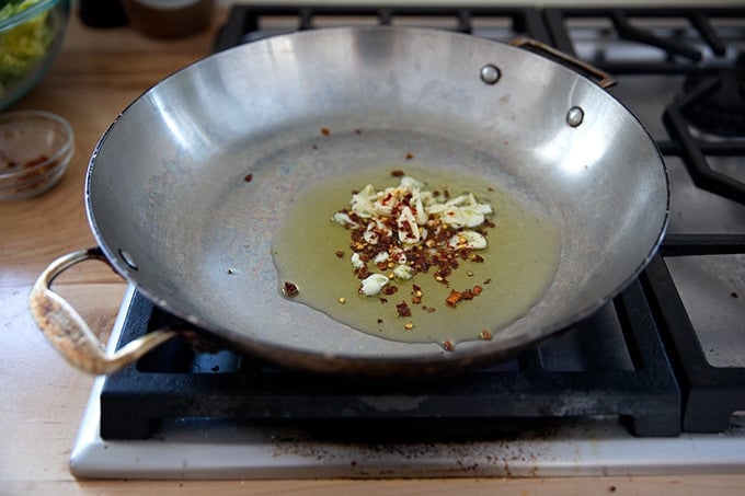 A large skillet stovetop with some olive oil, slice garlic, and crushed red pepper flakes inside.