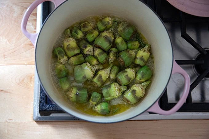 A large pot on the stovetop filled with baby artichokes cooking in olive oil, water, rosemary and garlic.