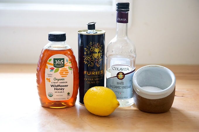 A bottle of honey, olive oil, white balsamic vinegar, salt and a lemon on a countertop.