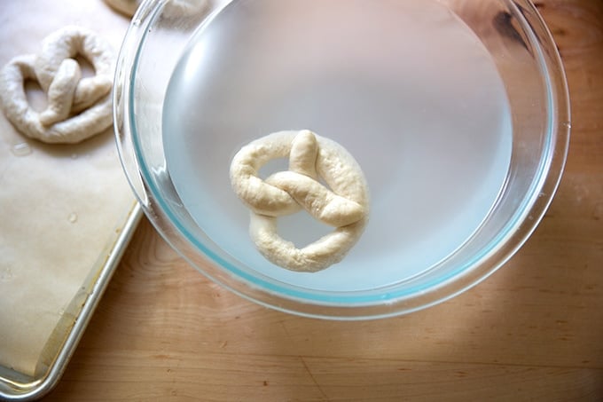 A shaped pretzel floating in a bowl of hot water with baking soda.