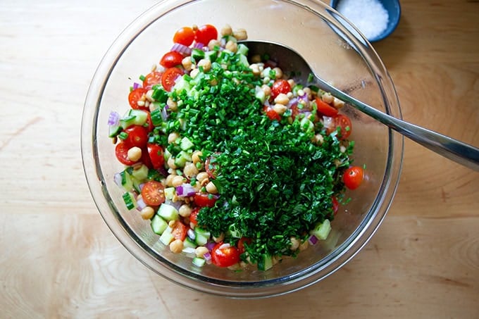 A large bowl filled with chickpeas, tomatoes, cucumber, red onion, toasted pine nuts, chopped chives, chopped parsley, and lemon vinaigrette.