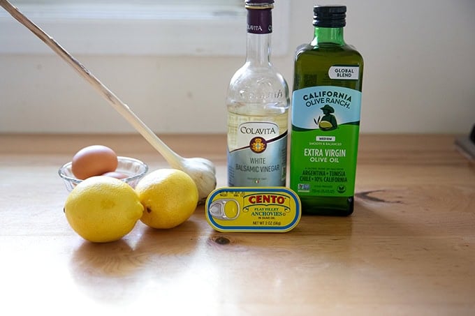 Ingredients to make Caesar dressing on a counter top: eggs, lemon, garlic, anchovies, white balsamic vinegar, and extra-virgin olive oil.