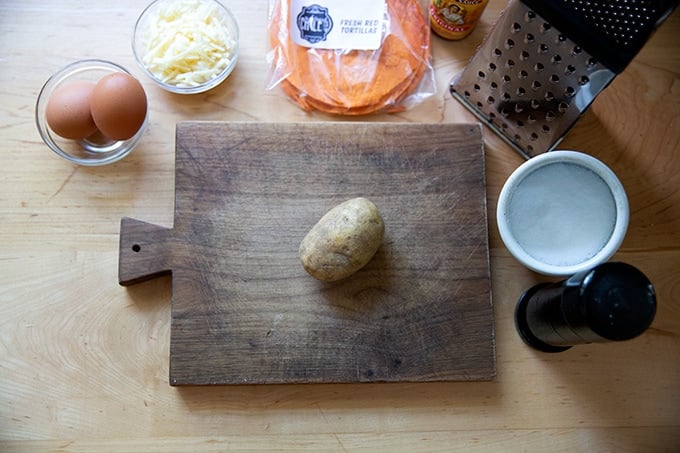 A cutting board with a potato on it aside eggs, cheese, tortillas, salt, pepper and a box grater.