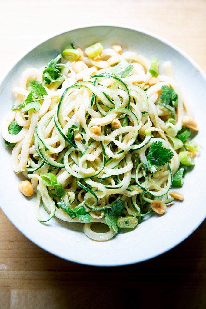 A bowl of peanut noodles with cucumbers, scallions and peanuts.