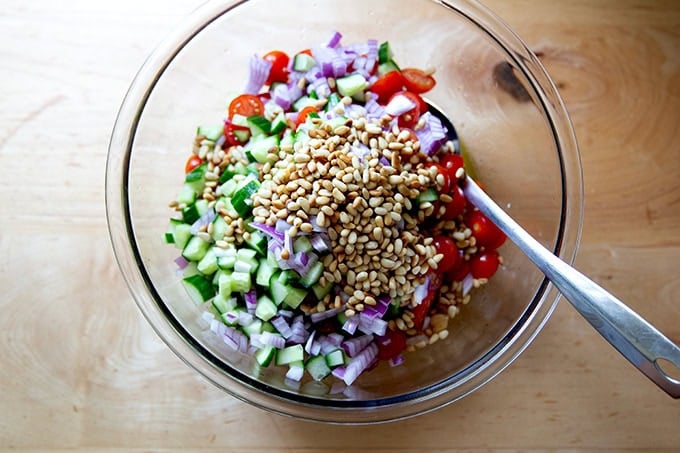 A large bowl filled with chickpeas, tomatoes, cucumber, red onion, toasted pine nuts, and lemon vinaigrette.