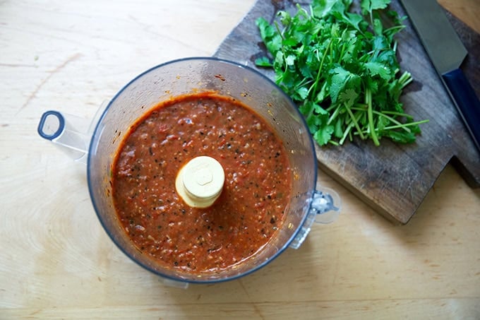 A food processor filled with puréed blistered tomato salsa aside a bundle of cilantro.
