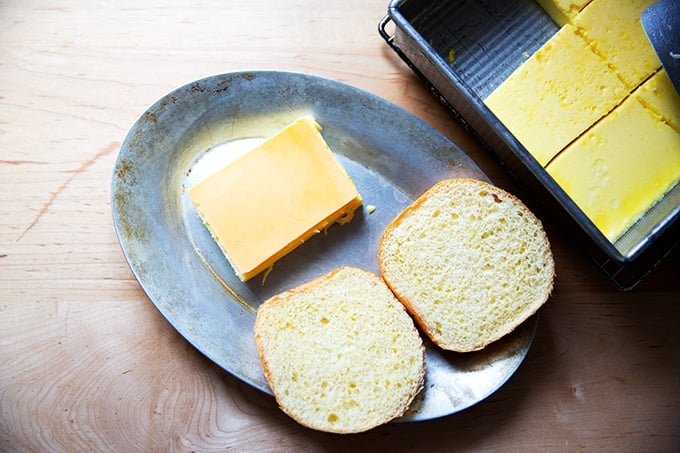 A halved brioche bun on a sizzle pan aside an egg slice topped with cheese.