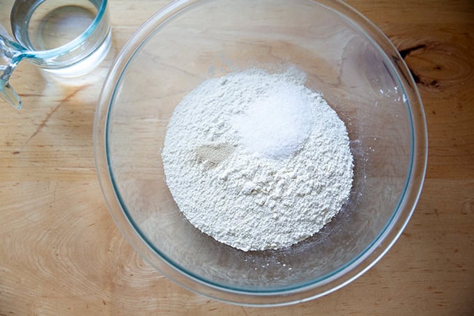 A large glass bowl filled with flour, salt, and instant yeast aside a liquid measure filled with water.