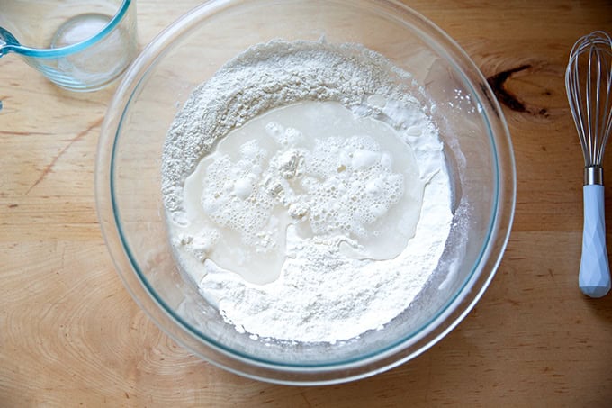 Water added to a large glass bowl filled with flour, salt, and instant yeast.