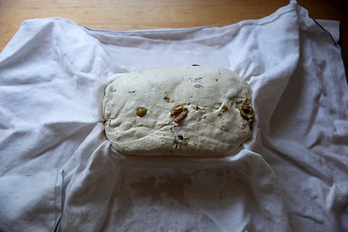 An unbaked loaf of rosemary-olive sourdough bread in a tea-towel lined banneton after 2 days in the fridge.