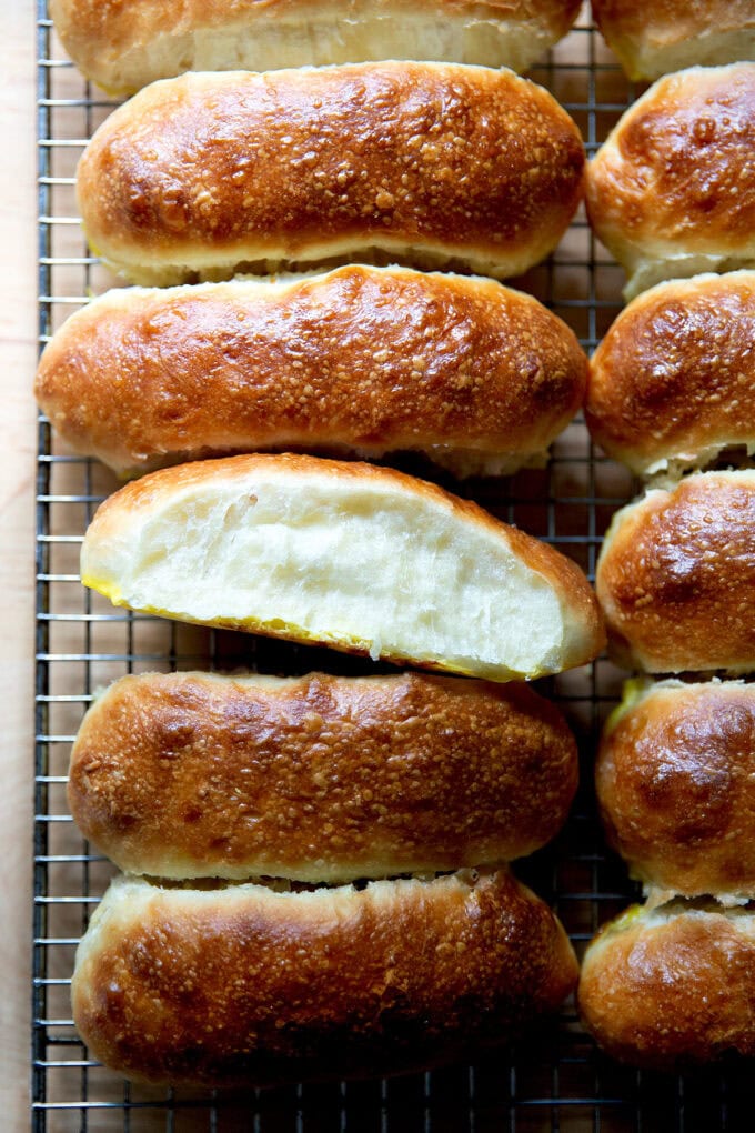Twelve just-baked brioche hot dog buns on a cooling rack with one turned on its side.