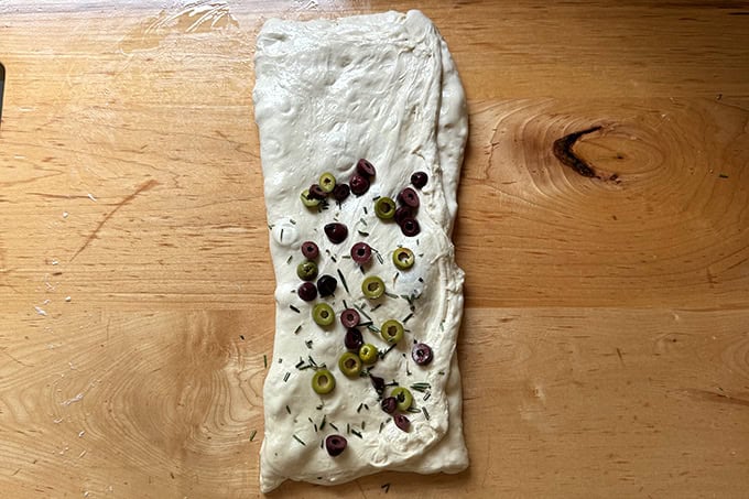 Rosemary-olive sourdough dough in the process of being folded on a countertop to incorporate the olives and rosemary.