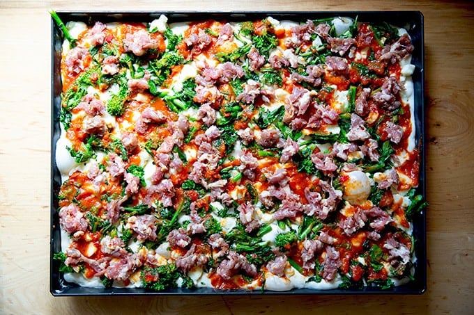 An unbaked grandma-style pizza topped with cheese, sauce, sautéed broccoli rabe and sausage.