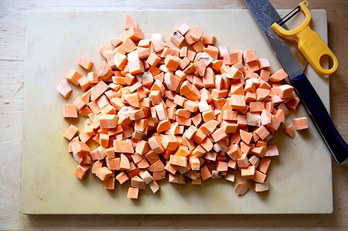 Cubed sweet potatoes on a cutting board aside a chefs knife and peeler.