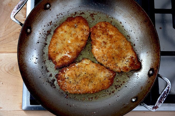 Three chicken cutlets frying in a large skillet on the stovetop.